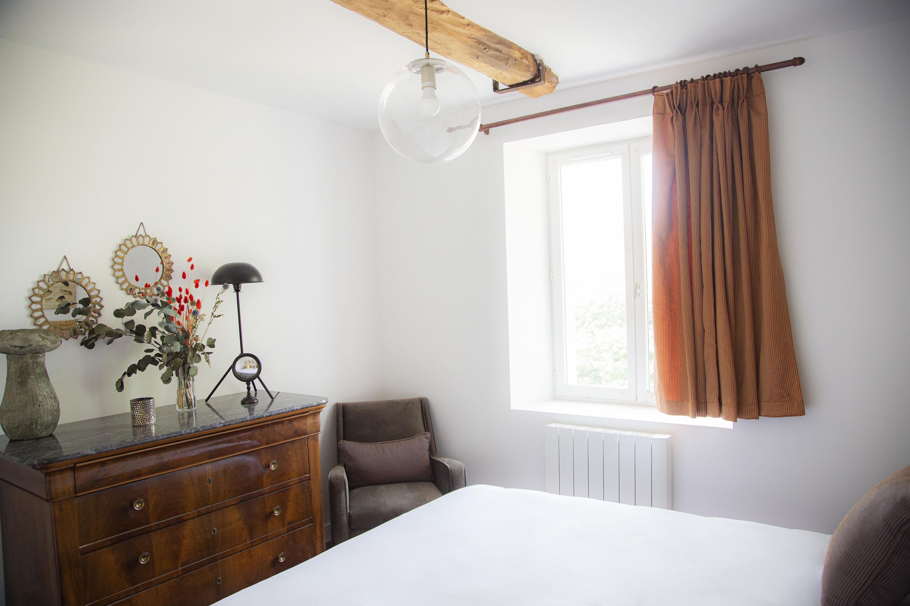 A bedroom with a wooden dresser, a chair, a bed, a window with curtains, and wall decor.