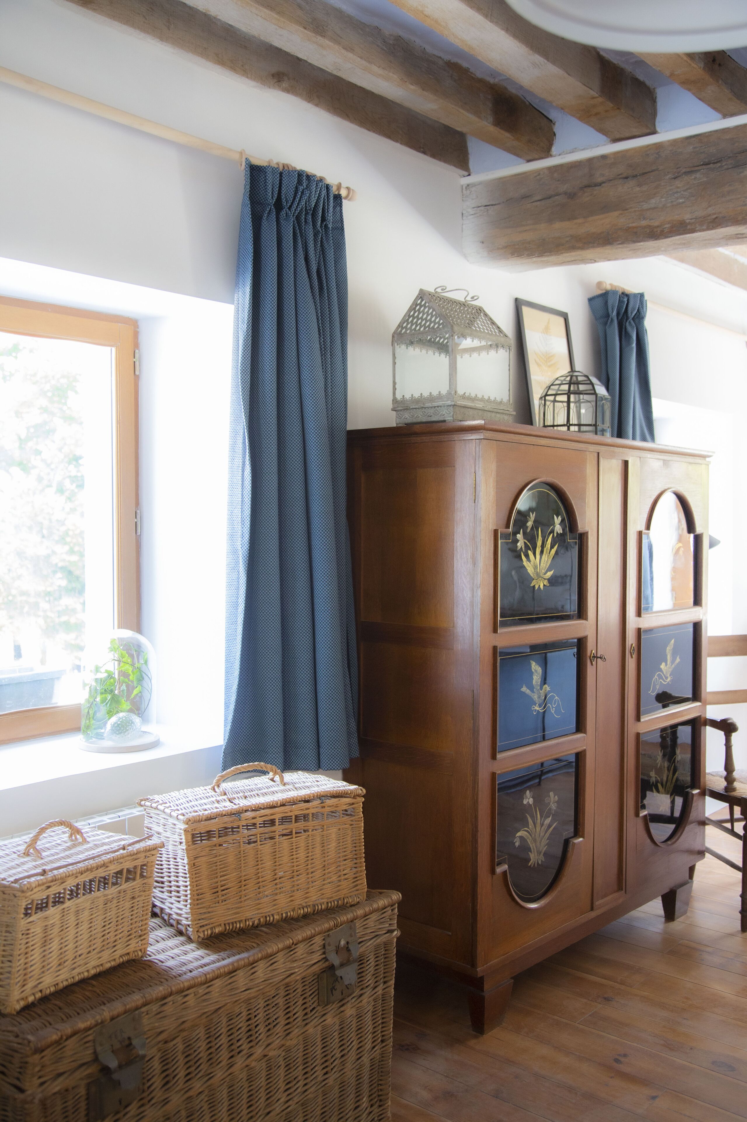 A wooden cabinet with glass doors, wicker baskets, and a window with blue curtains.