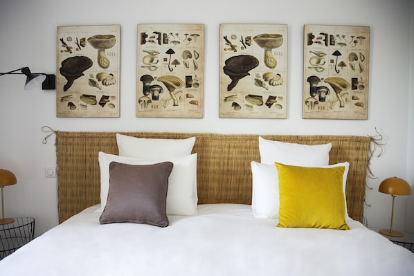 A neatly made bed with white and colored pillows, a woven headboard, and three botanical posters on the wall.