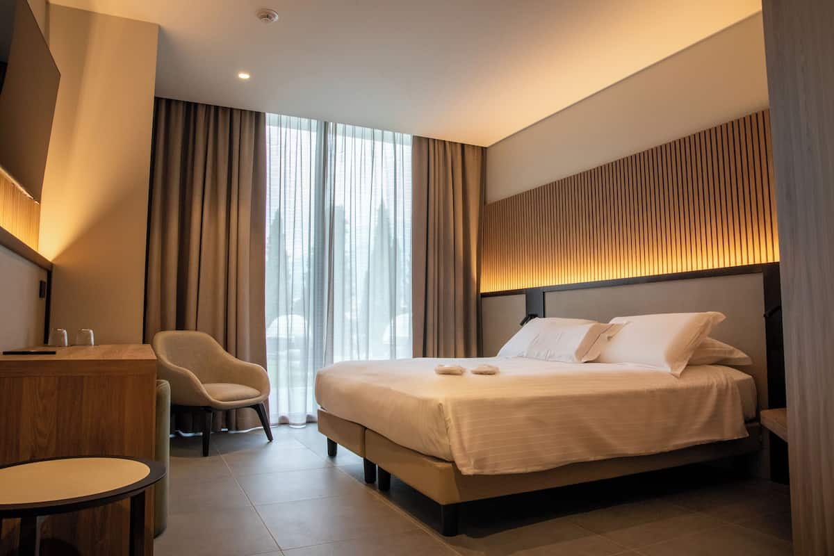 A modern hotel room with a large bed, a desk, a chair, and a view of the outdoors.