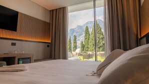 Habitación de hotel con una cama grande, un televisor de pantalla plana montado en la pared, un escritorio pequeño con una silla y vistas a las montañas a través de amplios ventanales.