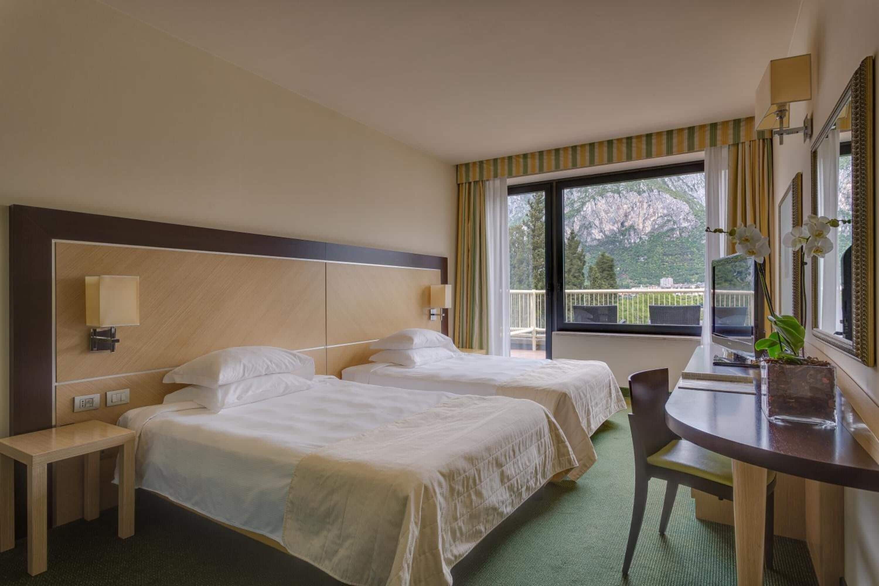 Een hotelkamer met twee bedden, een bureau, een stoel, een raam met gordijnen en uitzicht op groen.