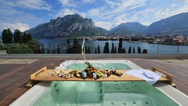 Un área de piscina en la azotea con terraza de madera, jacuzzi, y una mesa dispuesta con comida y bebidas, con vista a un lago y montañas.