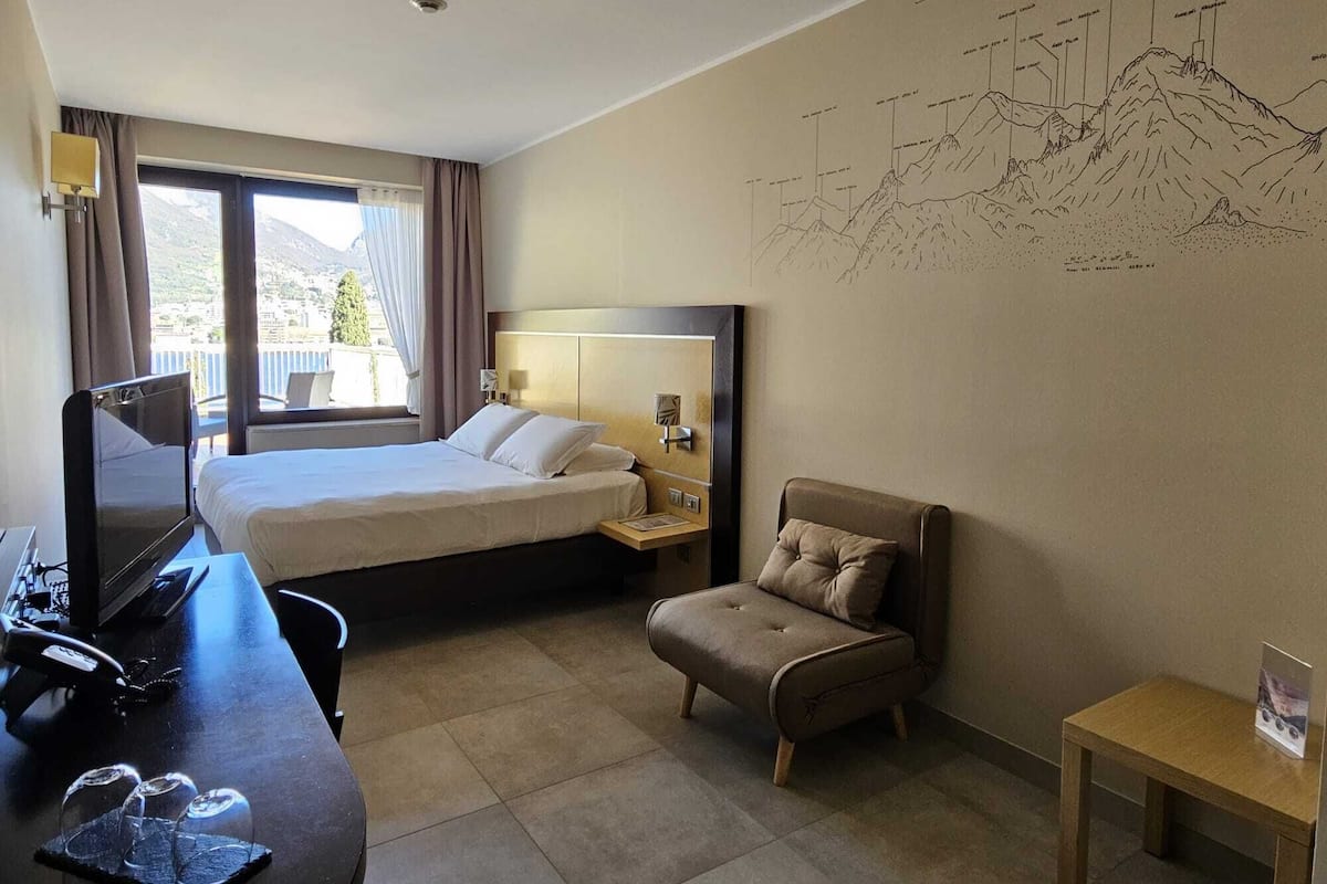 A hotel room with a bed, a desk with a computer, a chair, and a wall with a mountain drawing.
