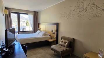 A hotel room with a bed, a desk with a computer, a chair, and a wall with a mountain drawing.