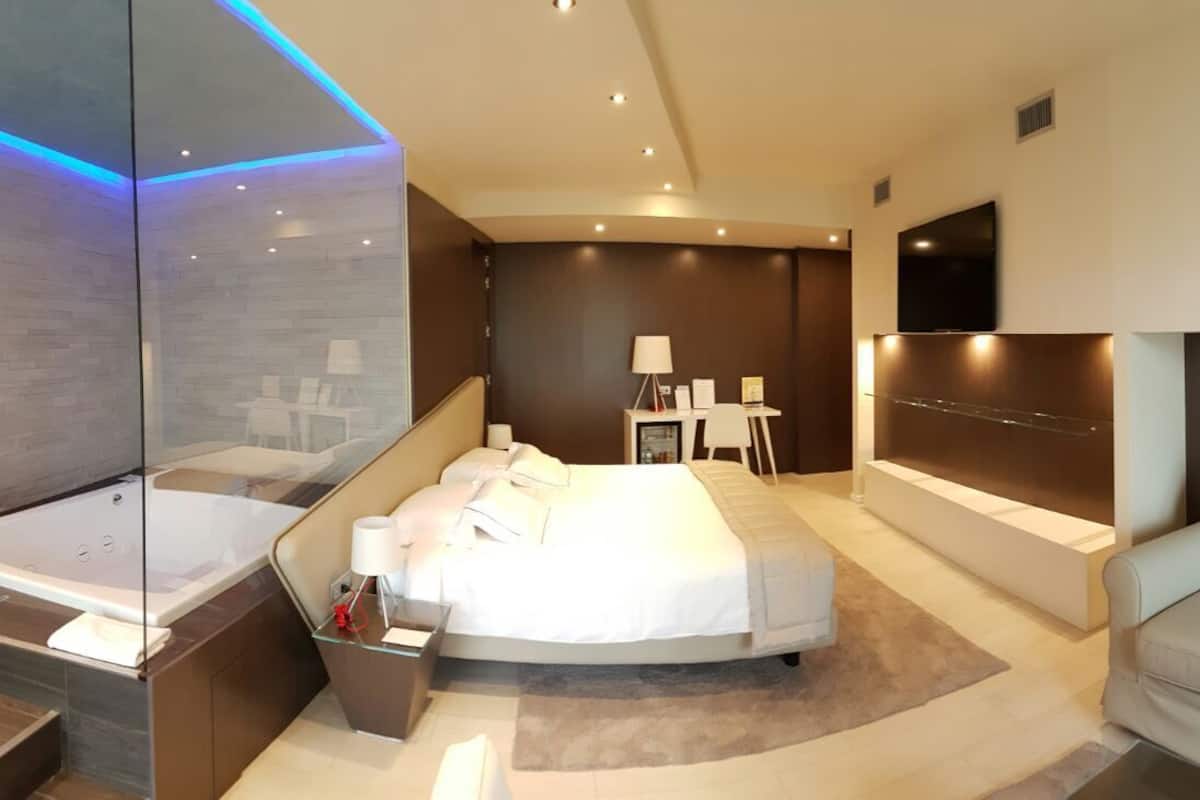 A modern hotel room with a large bed, a sitting area, a bathtub, and a TV mounted on the wall.