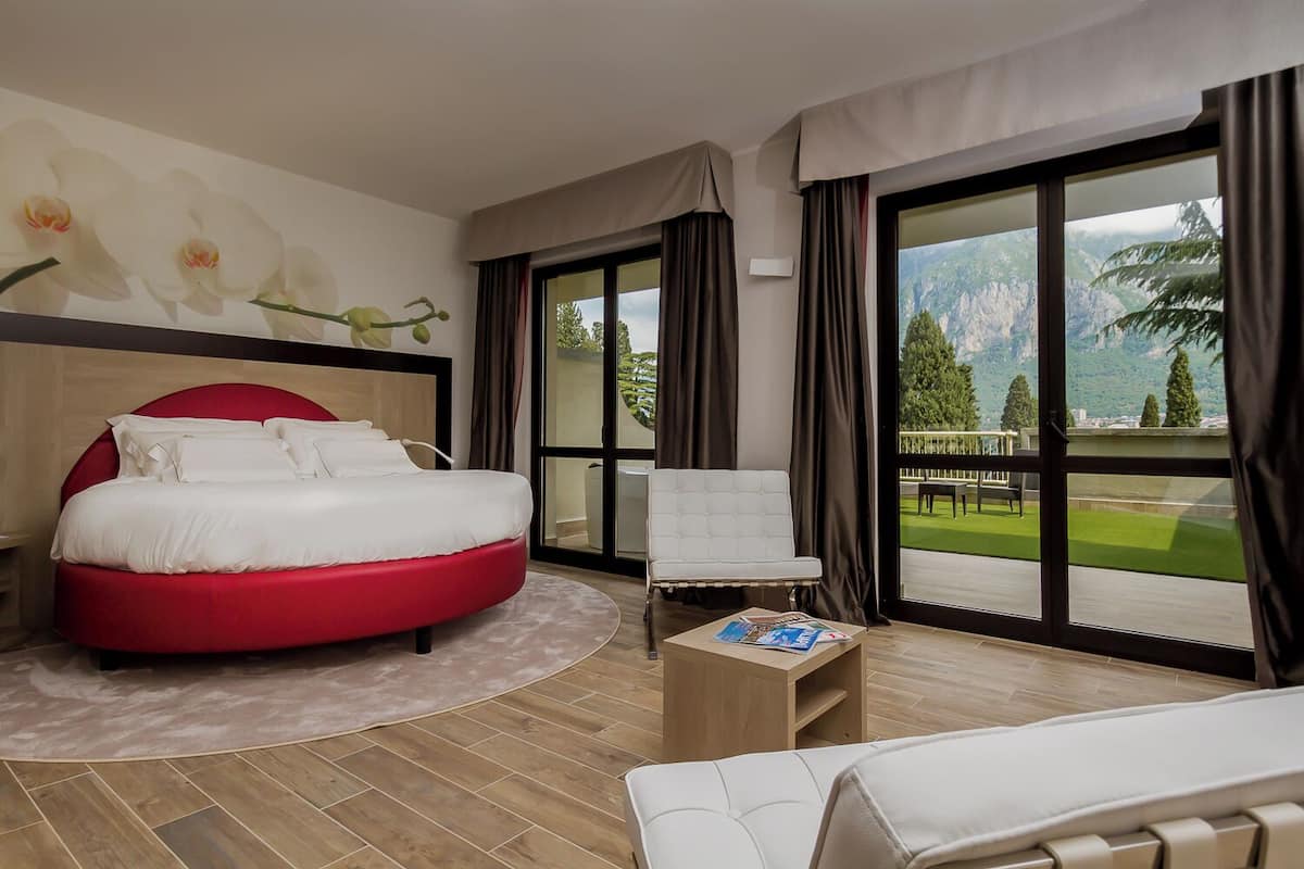 A modern hotel room with a red bed, a white armchair, a small wooden table, and a large window offering a mountain view.