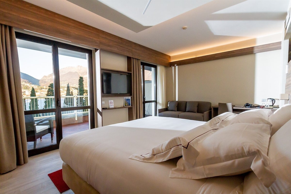 A hotel room with a large bed, a TV, a sofa, and a balcony with a mountain view.