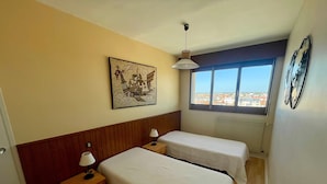 Habitación de hotel con dos camas, cabecera de madera, mesita de noche con lámpara, ventana con vistas a la ciudad y una obra de arte colgada en la pared.