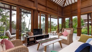 Een ruime woonkamer met rieten meubilair, een flatscreen televisie en een groot raam met uitzicht op een tropische tuin.