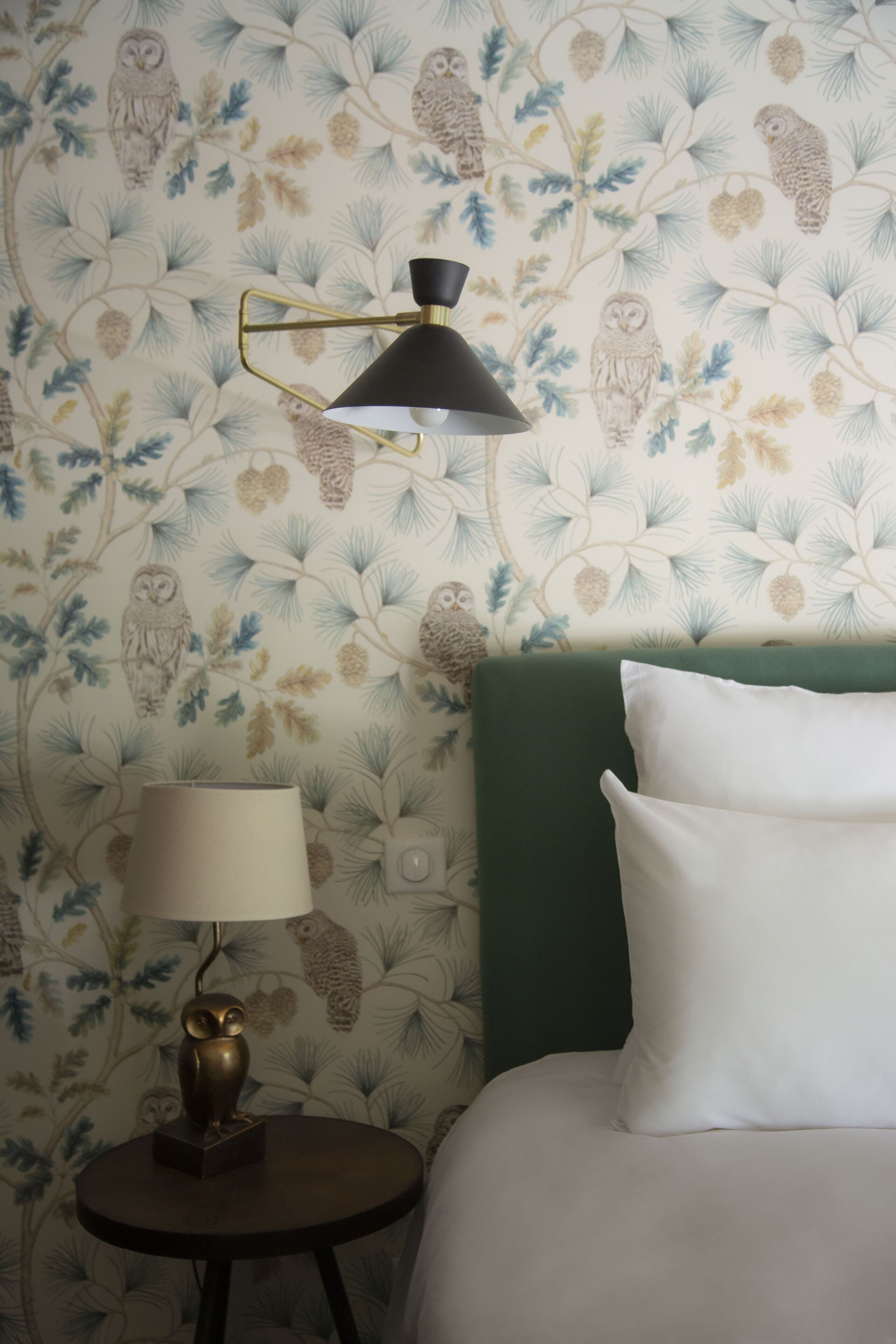 Et værelse med en seng, et rundt bord med en lampe, og en væg med blomstret tapet med ugler.