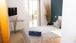 Une chambre à coucher avec un lit, un téléviseur, un radiateur et une vue sur l’extérieur, visible à travers une porte.