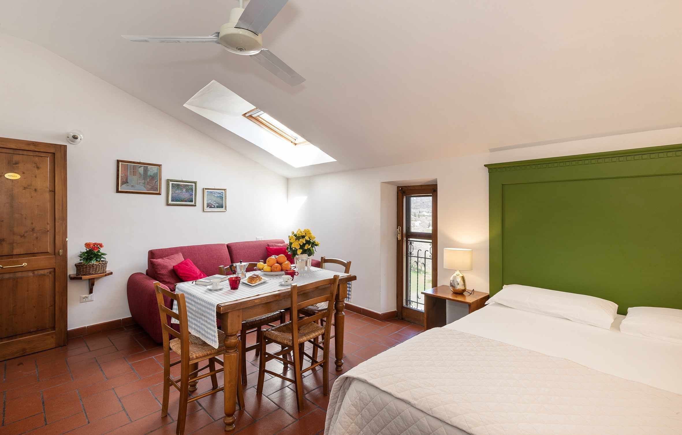 A bedroom with a green headboard, a dining area with a table and chairs, a red sofa, and a ceiling fan.