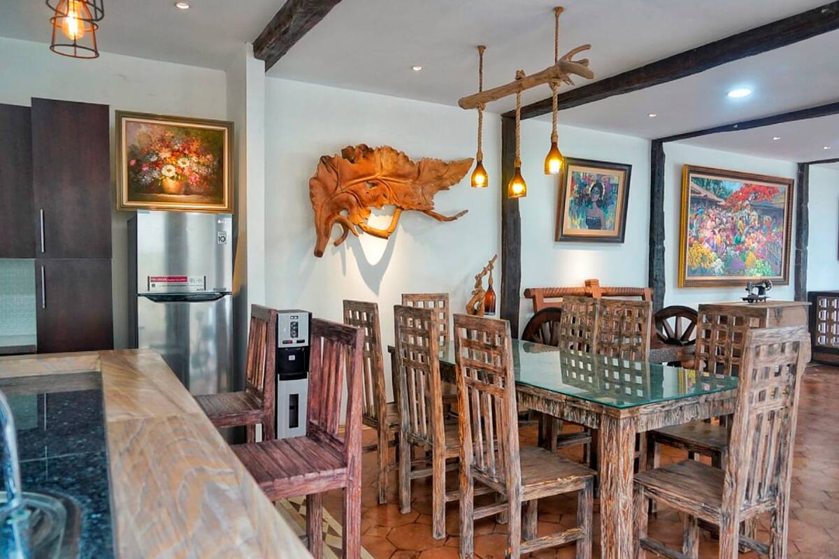 A dining area with a glass-top table, wooden chairs, and a rustic decor including a large leaf sculpture and hanging lights.