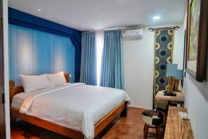 A hotel room with a wooden bed, blue curtains, an air conditioning unit, a patterned curtain, a small table with a lamp, and a stool. - Villa Himala Bali (Seminyak)
