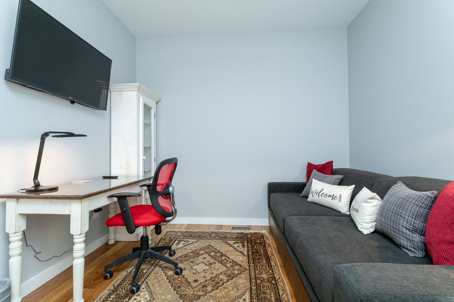 A modern living room with a grey sofa, a red office chair, a desk with a lamp, and a flat-screen TV mounted on the wall.