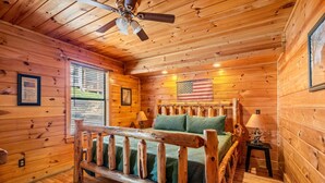 Interior de una cabaña de madera con una cama, un ventilador de techo y una bandera estadounidense en la pared.