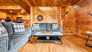 Interior de una cabaña con paredes de madera, un sofá, una mesa de centro, una escalera y una zona de cocina.