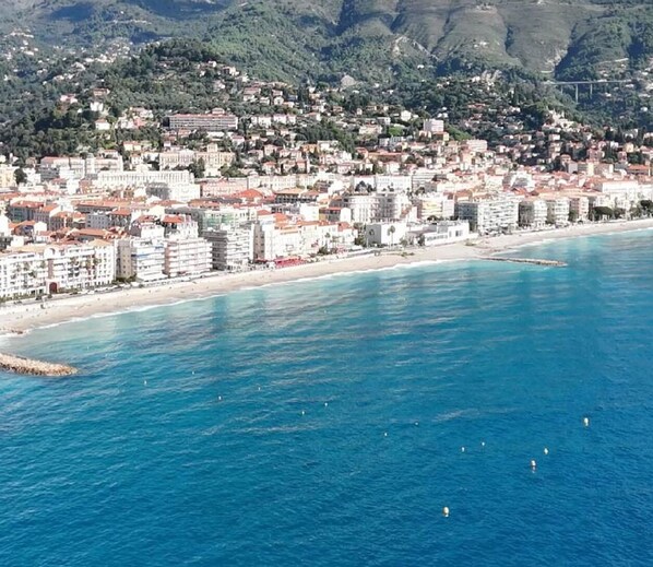 A coastal cityscape with buildings along the waterfront, a clear blue sea, and mountains in the background. - Stunning 1-Bedroom Apt with Sea View, Terrace, and Private Parking - Menton, Garavan Area (Menton)