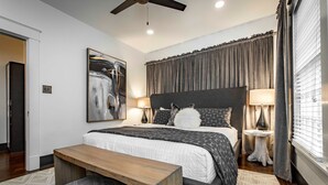A modern bedroom with a large bed, a wooden bench, a ceiling fan, and a painting on the wall. - Hip and Historic Home Awaits You!  Stroll downtown/Steps away from UCA and CBC (Conway)