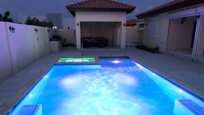 Une piscine rectangulaire bordée d’une terrasse carrelée, un espace de détente couvert et, au fond, un bâtiment avec un toit recouvert de tuiles rouges.