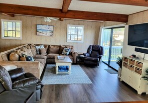 A living room with a sofa, armchairs, a coffee table, and a TV. There are windows and a balcony visible.