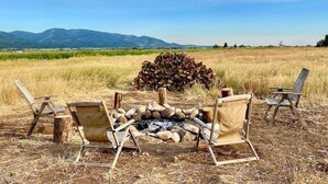 Uma área de estar rústica ao ar livre, com lareira rodeada por cadeiras, com vista para um campo e montanhas.