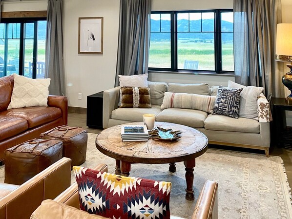 A living room with a brown leather sofa, a beige sofa with patterned cushions, a round wooden coffee table with books and a decorative bowl, and a view of a green landscape through large windows.
