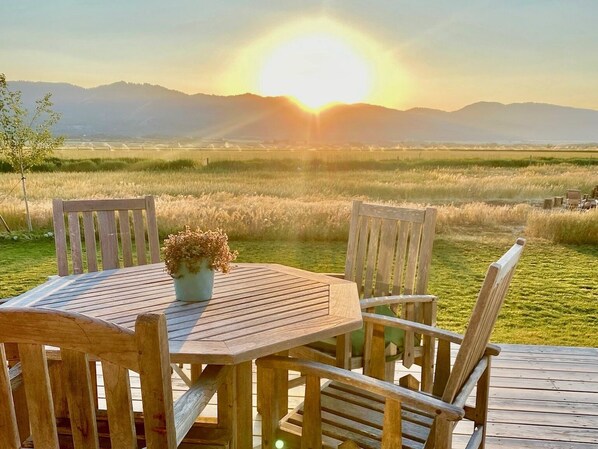 A wooden deck with a table and chairs, a potted plant, and a view of a sunset over a field and mountains.