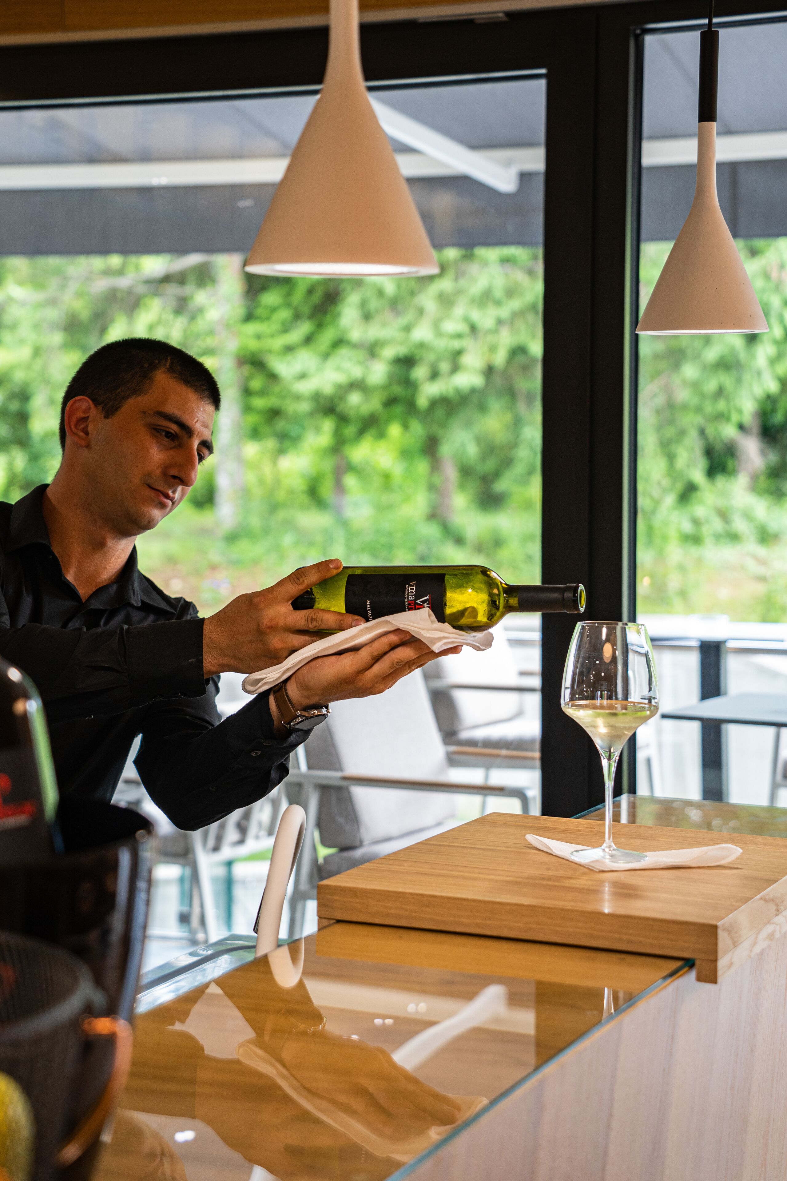 Une personne verse du vin dans un verre au comptoir d’un bar, avec vue sur la verdure à l’extérieur.
