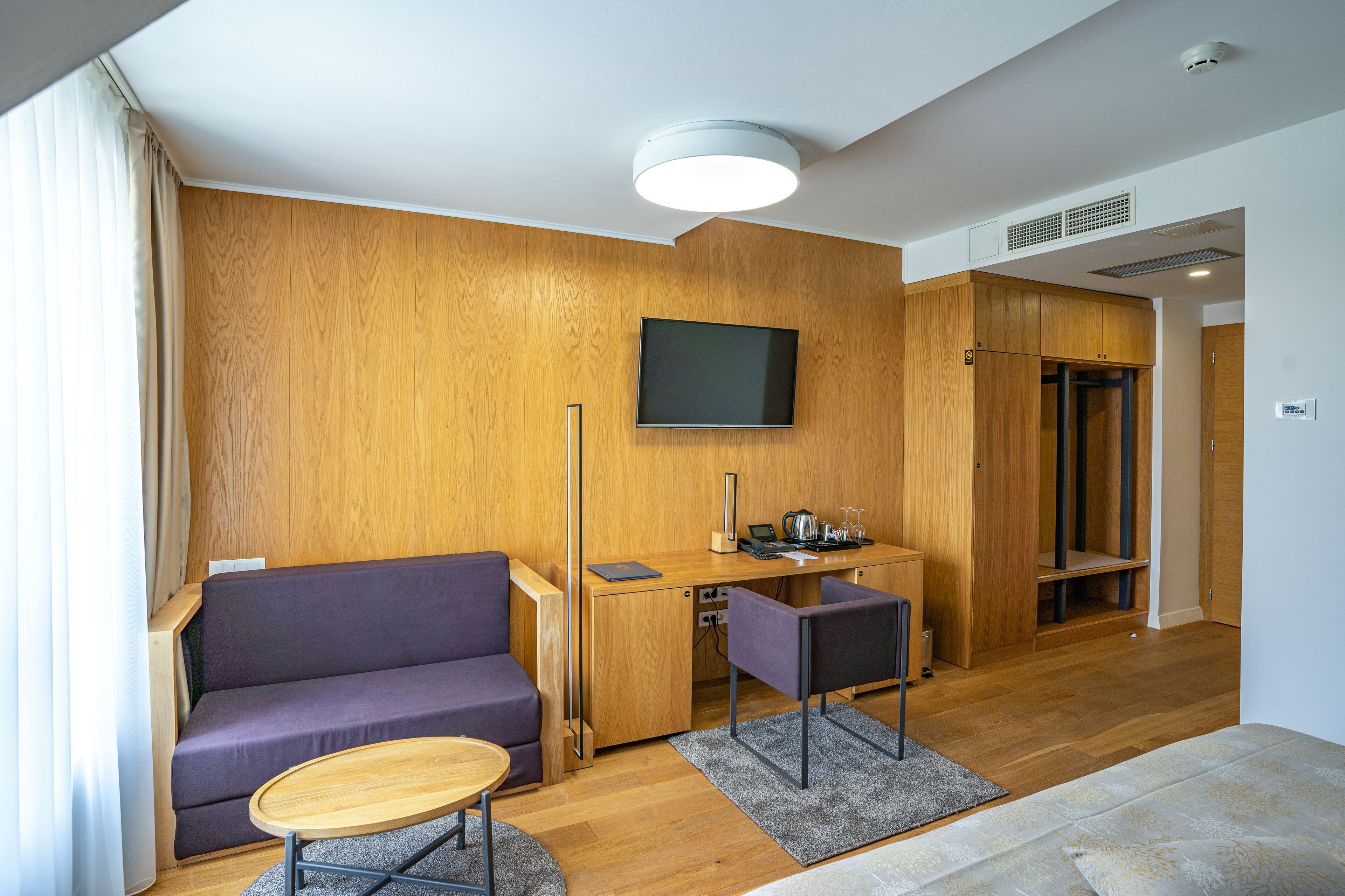 A modern room with a wooden wall, a sofa, a desk with a chair, a TV mounted on the wall, and a door leading to another room.