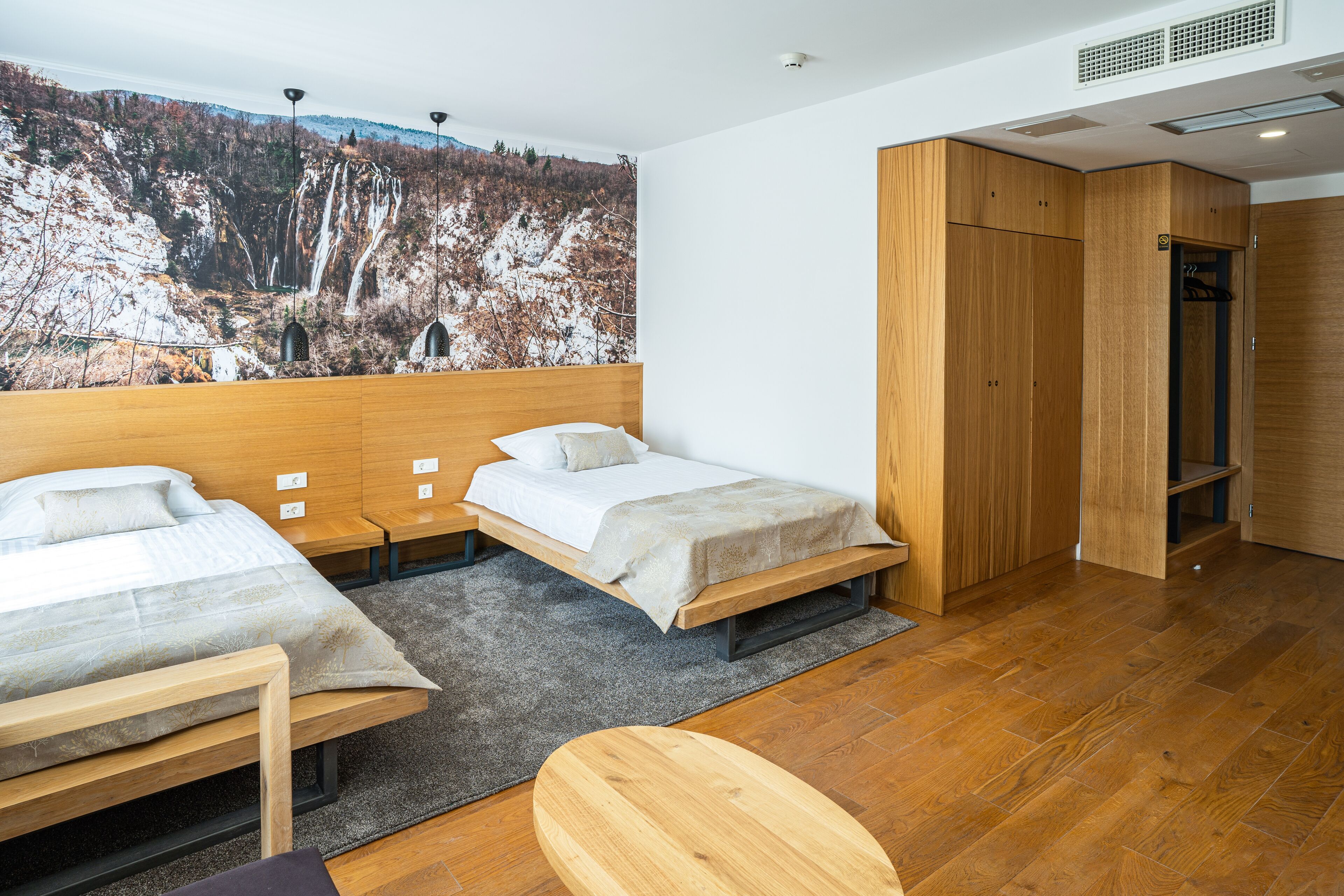 A modern hotel room with two beds, a wooden wardrobe, and a large wall mural of a mountainous landscape.