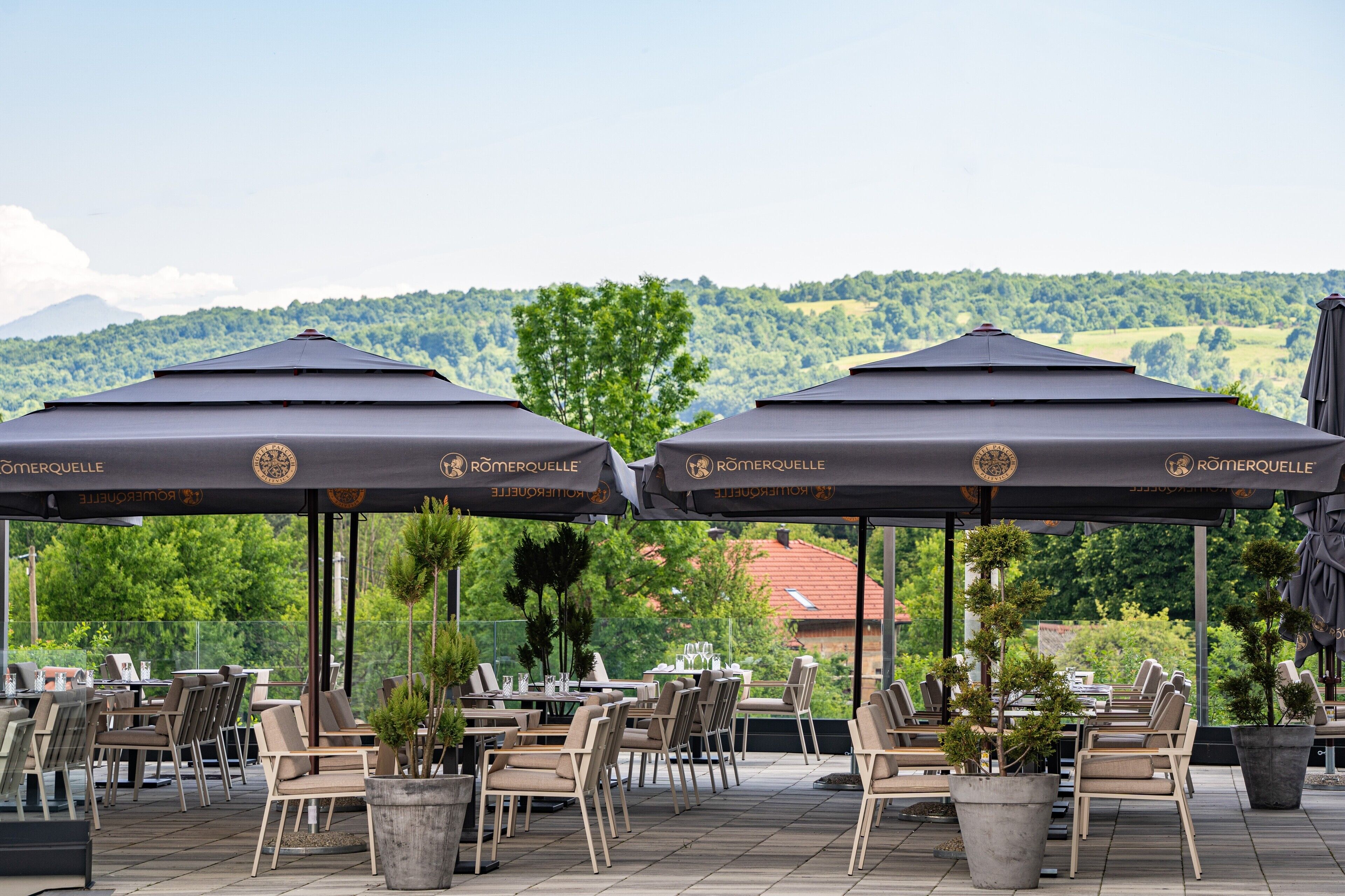 An outdoor patio with tables and chairs under umbrellas, a view of a green landscape, and a building in the background.