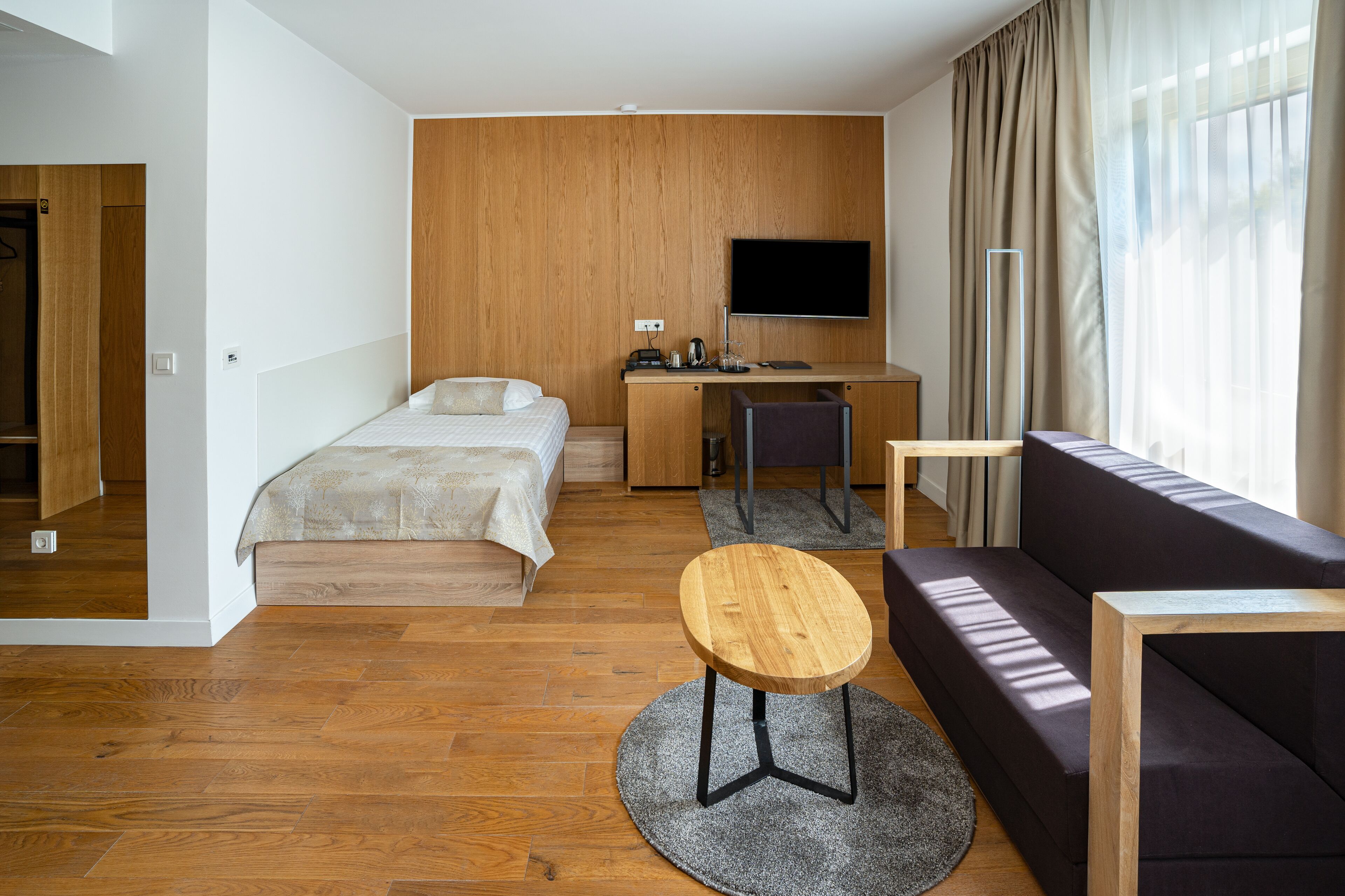 Habitació d'hotel moderna amb un llit, una taula de treball amb televisió de pantalla plana, un sofà i una taula rodona petita.