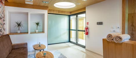Un área de recepción con un mostrador de madera, un sofá marrón, dos mesas redondas con taburetes y una puerta de cristal que da a otra habitación.