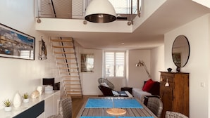 A spacious living area with a high ceiling, a staircase, a dining table with blue placemats, wicker furniture, and a large mirror. - ❤️ New! Luxury apartment with sea view in the heart of downtown (Concarneau)