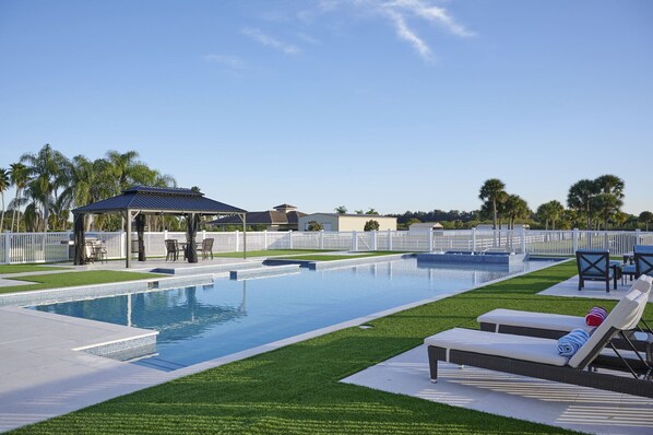 Una piscina con solárium, sillas de descanso y una zona de estar cubierta.