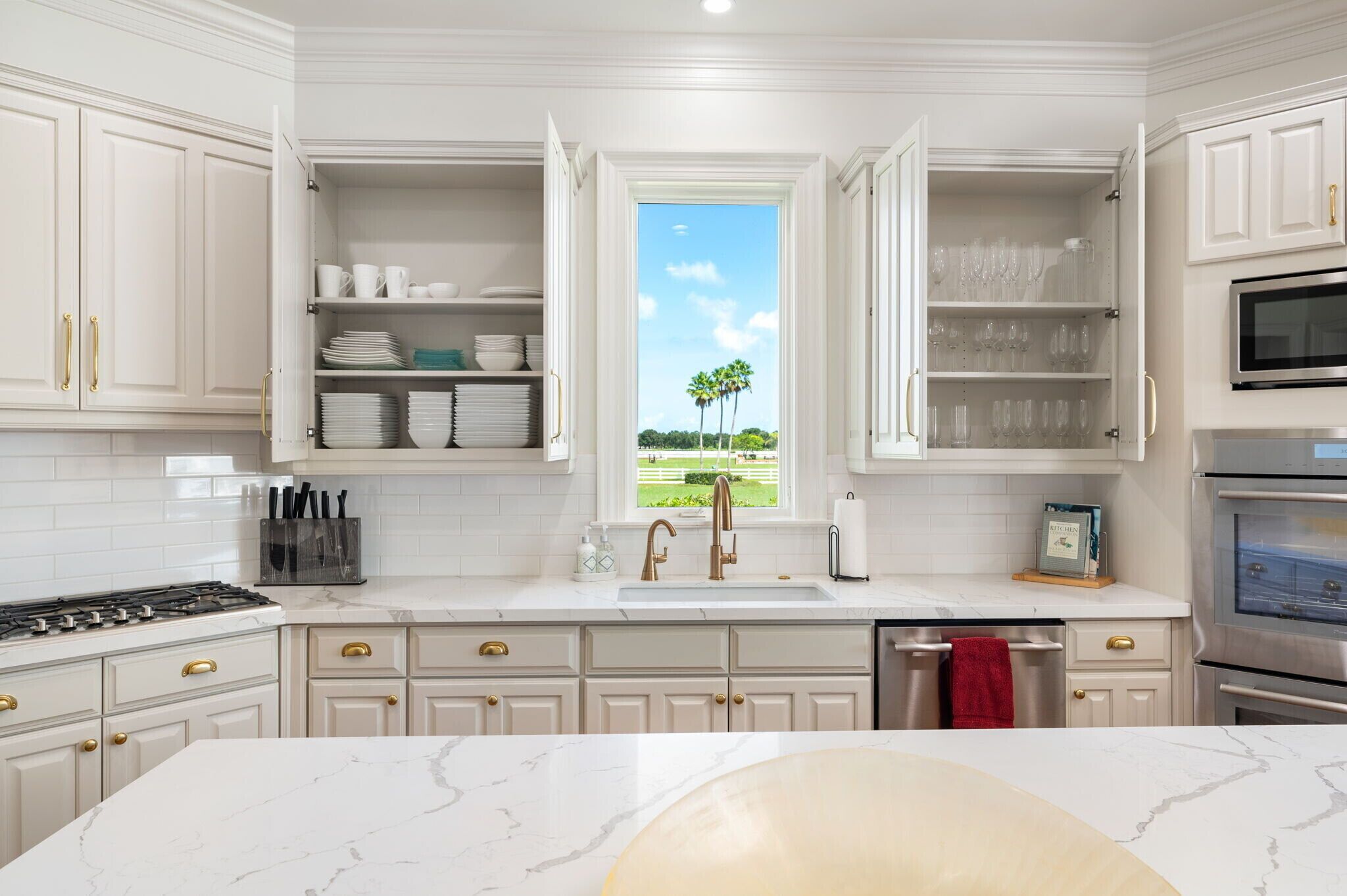 Una cocina con armarios blancos, estufa a gas, lavamanos y ventana con vista a palmeras.