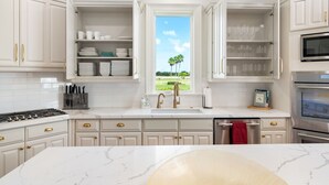 Una cocina con armarios blancos, estufa a gas, lavamanos y ventana con vista a palmeras.