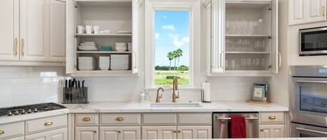 Una cocina con armarios blancos, estufa a gas, lavamanos y ventana con vista a palmeras.