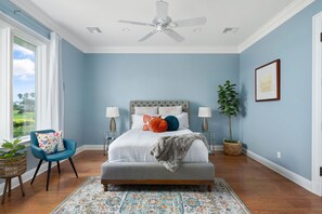 Un dormitorio con una cama grande, un ventilador de techo, una ventana con cortinas, una silla, una mesita de noche y una planta en maceta.