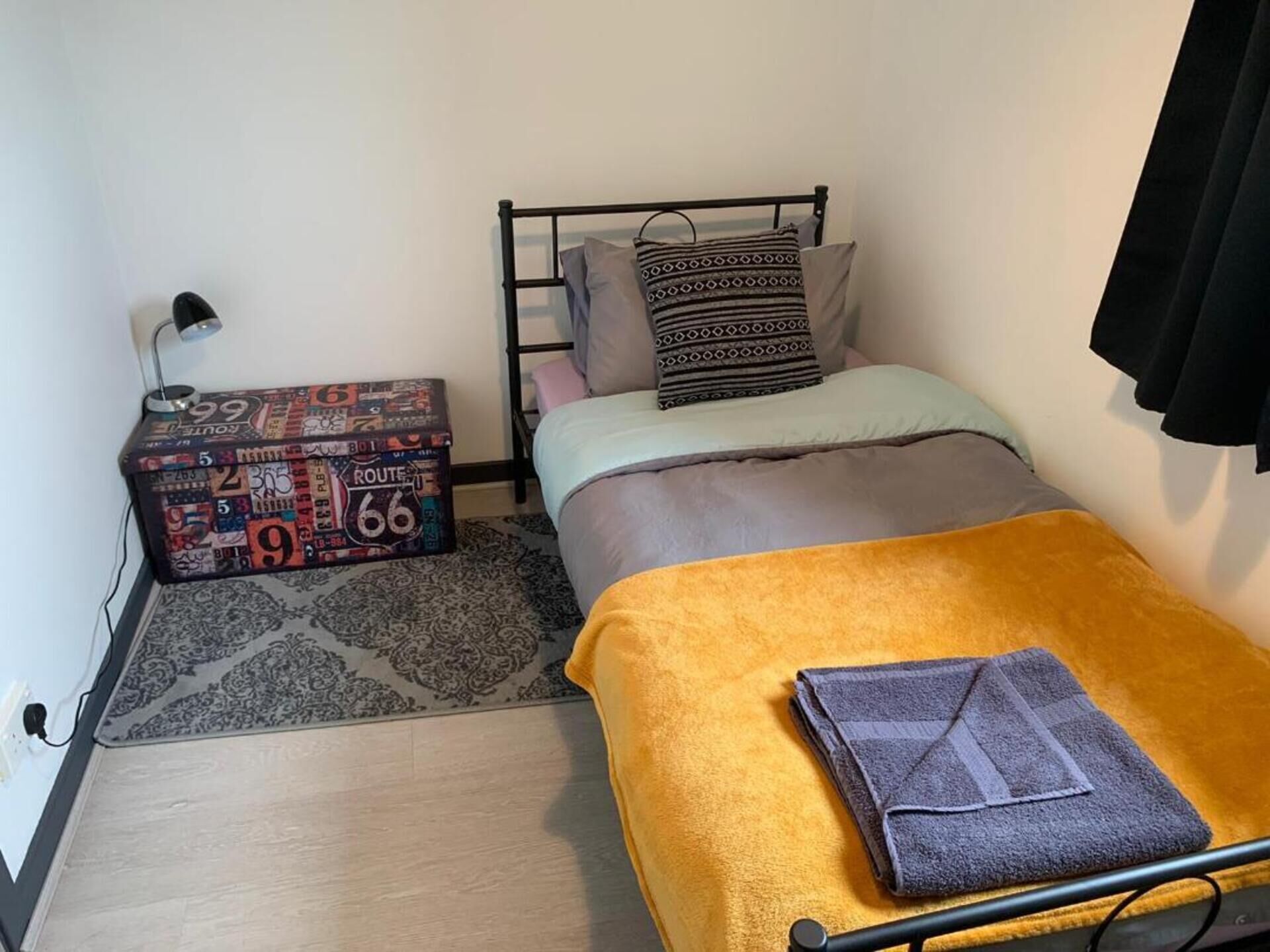 Une petite pièce, bien rangée, avec un lit, une table de chevet dotée d’une lampe et une boîte décorative.