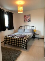 Un dormitorio con una colcha a cuadros, un cabecero de metal negro, una mesita de noche pequeña con una lámpara amarilla y una alfombra mullida.
