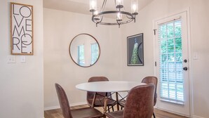 A dining area with a round table, four chairs, a chandelier, a mirror, and a door with blinds.