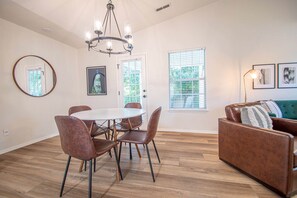 Uma sala de jantar com mesa redonda e cadeiras, um lustre e um sofá com almofada.