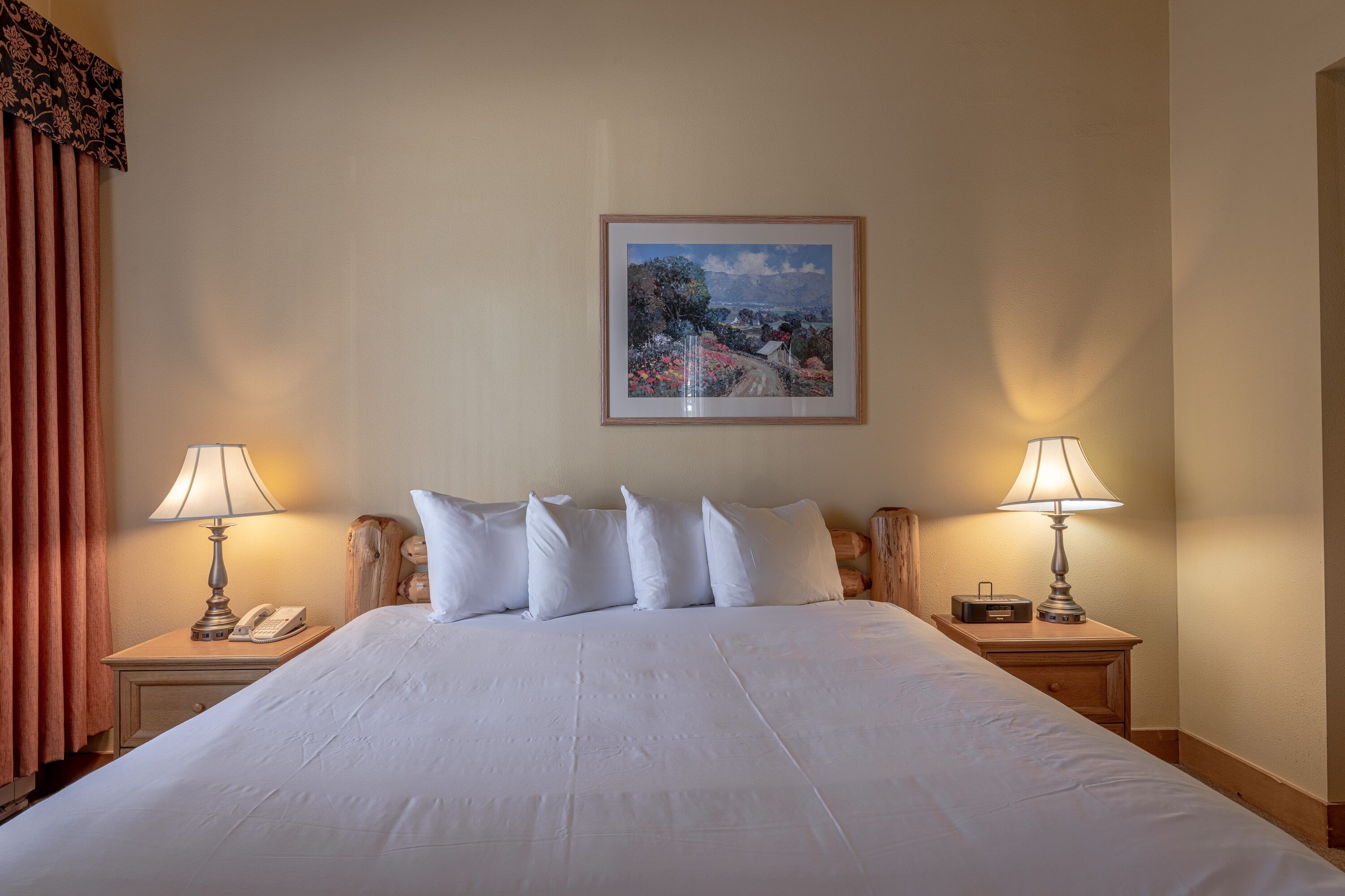A neatly made bed with white linens, two bedside lamps, a telephone, and a framed landscape painting on the wall.
