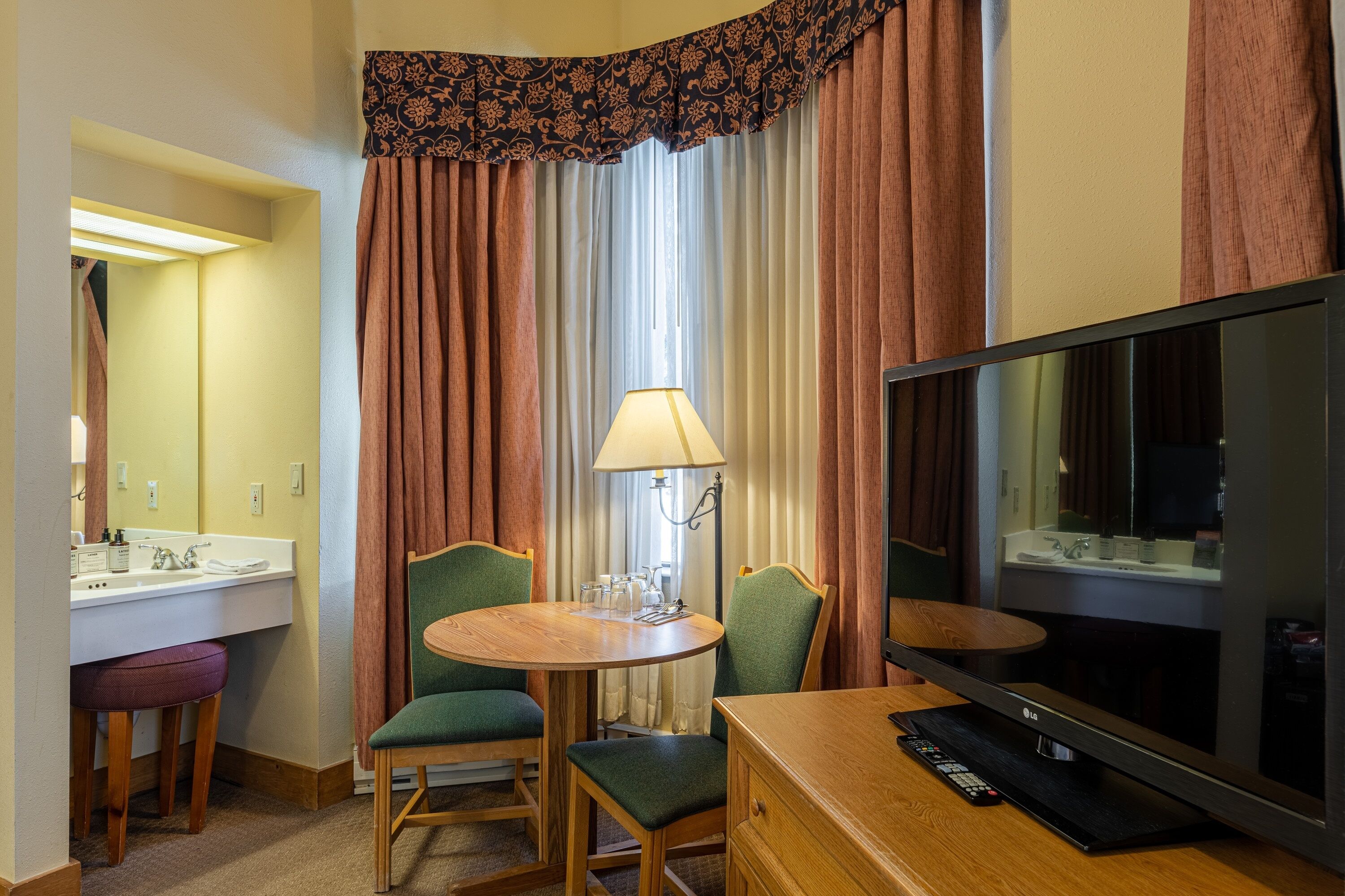 A hotel room with a TV, a small round table with chairs, a lamp, a mirror, and a bathroom with a sink and toilet.