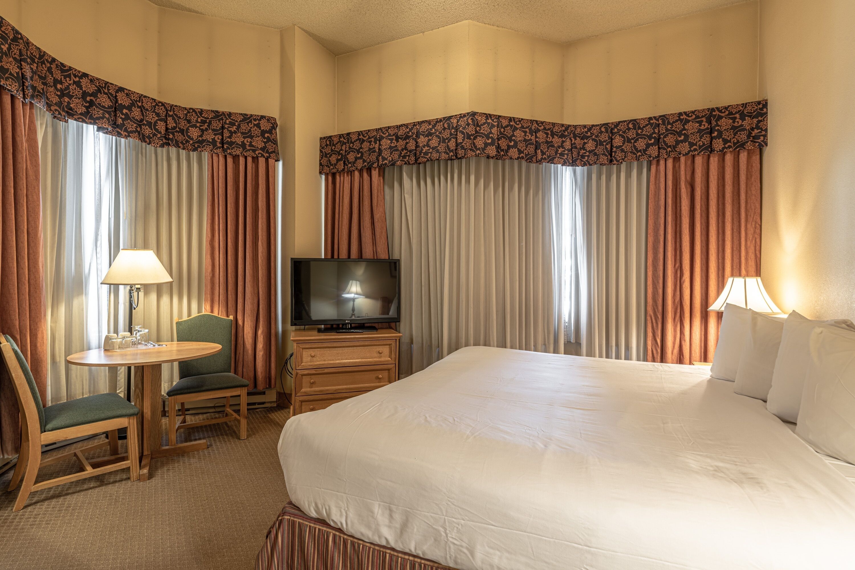 Habitación de hotel con una cama grande, un televisor sobre un soporte de madera, una mesita con una silla y cortinas con un frente decorado.