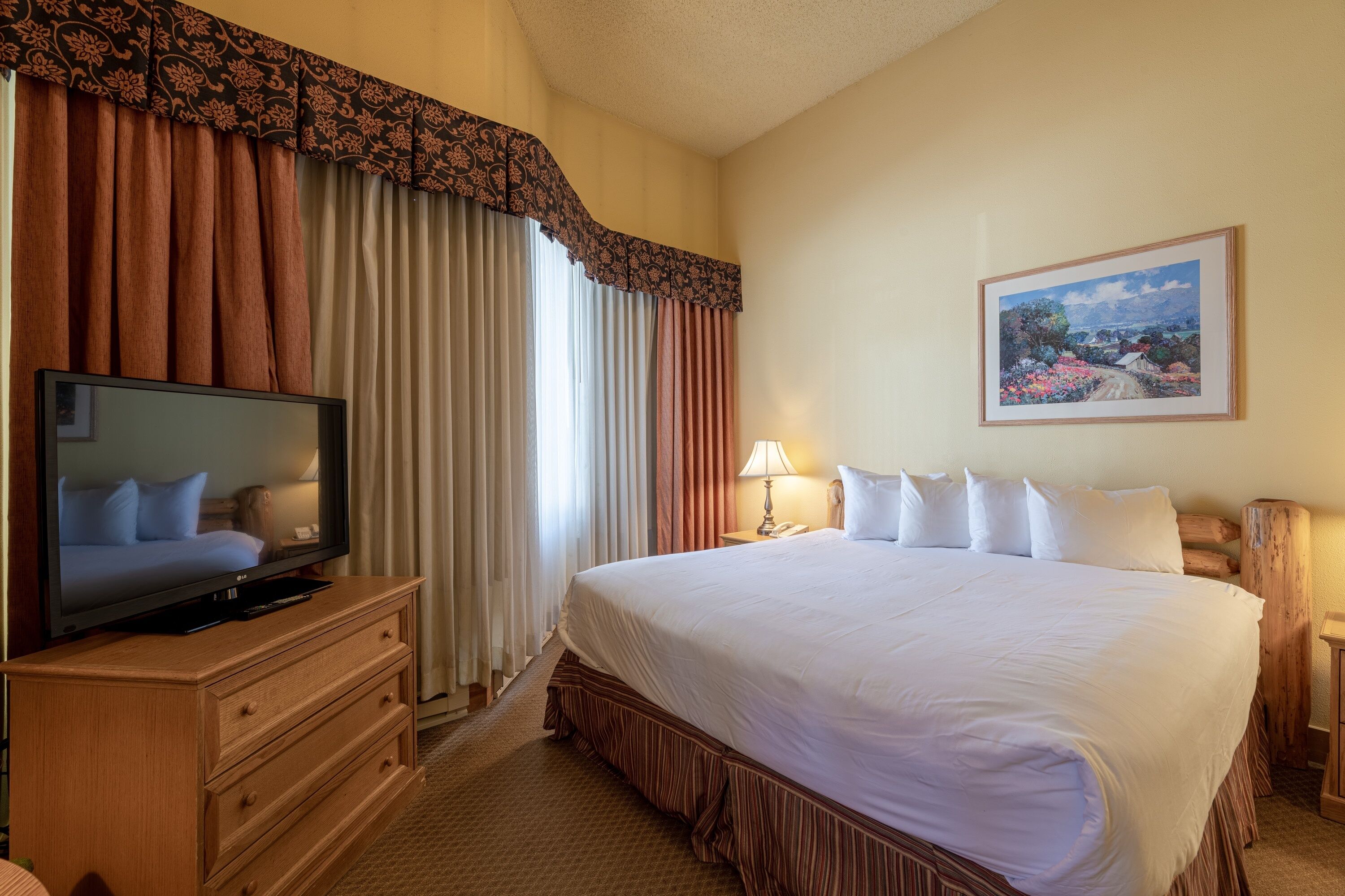 A hotel room with a large bed, a television on a wooden dresser, a painting on the wall, and a window with curtains.