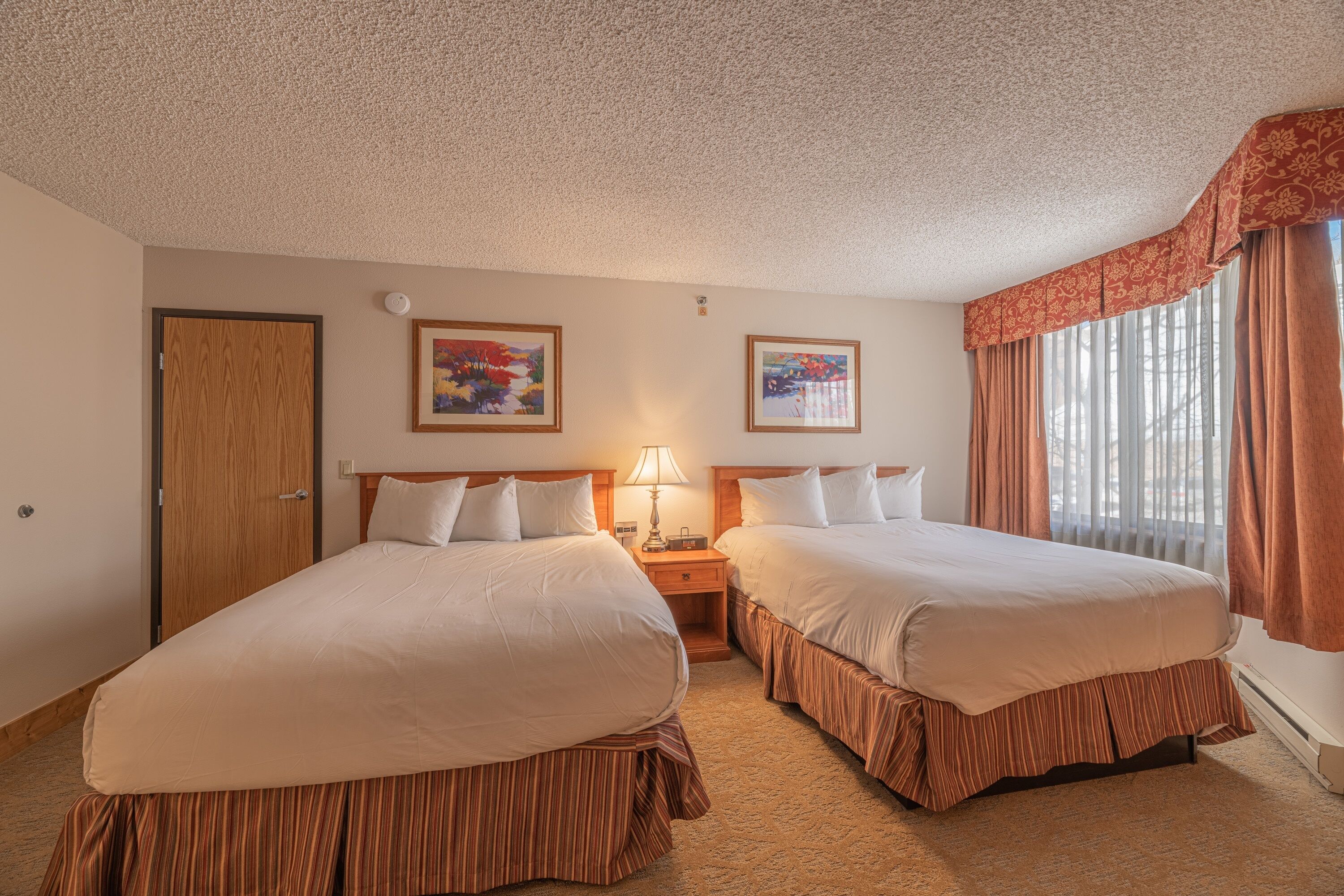 A hotel room with two beds, a wooden door, framed artwork on the walls, and a window with curtains.
